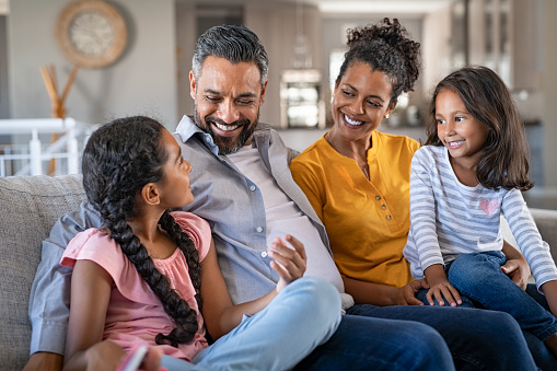 Cheerful multiethnic family at home sitting on sofa listening to daughter. Happy couple with two girls relaxing at home together. Smiling indian parents talking to their daughter at home in the living room.