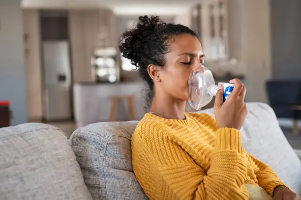 Black young woman holding a mask nebulizer inhaling fumes medication into lungs. African sick lady inhaling through inhaler mask sitting on the couch with copy space. Self treatment of the respiratory tract using inhalation nebulizer.