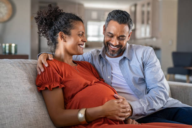 Happy mixed race couple expecting baby Mid adult couple expecting a baby while husband caresses the belly of his pregnant african woman. Happy father hands on expecting mother's baby bump while embracing her on couch. Lovely indian man touching belly of his girlfriend and feeling baby movement. human fertility stock pictures, royalty-free photos & images