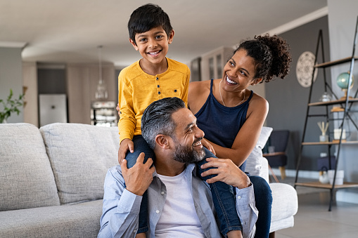 Cheerful african mother and indian father playing with son at home. Cute boy enjoying sitting on father shoulder while looking at camera. Middle eastern family having fun together on the sofa at home.