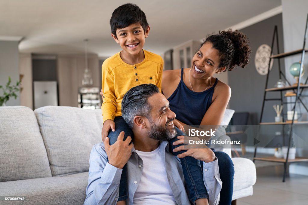 Happy ethnic family playing together at home Cheerful african mother and indian father playing with son at home. Cute boy enjoying sitting on father shoulder while looking at camera. Middle eastern family having fun together on the sofa at home. Family Stock Photo