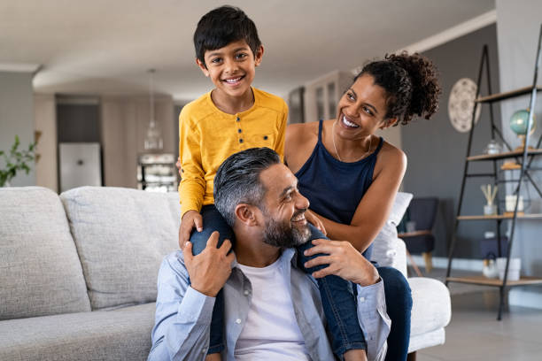 Happy ethnic family playing together at home Cheerful african mother and indian father playing with son at home. Cute boy enjoying sitting on father shoulder while looking at camera. Middle eastern family having fun together on the sofa at home. young family stock pictures, royalty-free photos & images