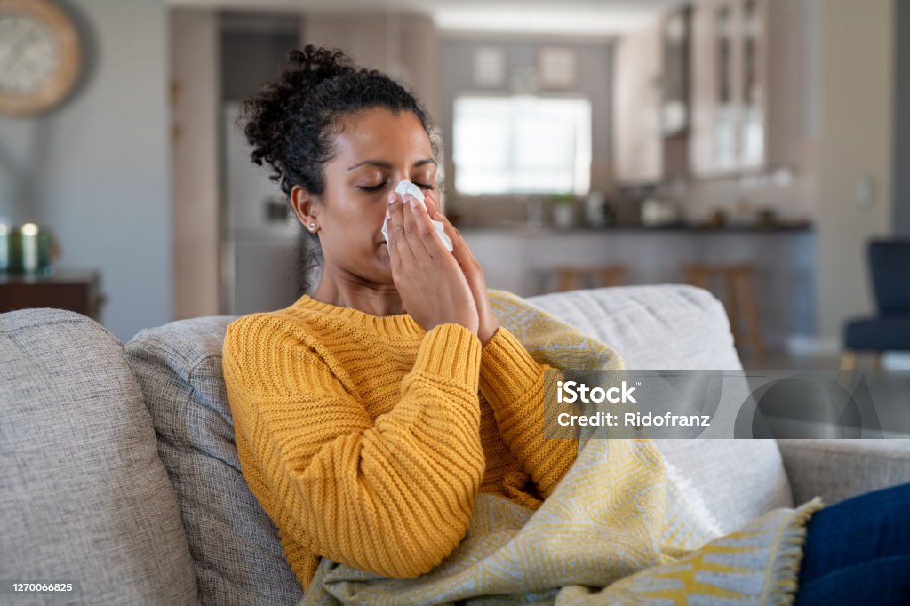 Cold sick african woman blowing nose Portrait of young black woman sneezing in to tissue at home. Sick african woman wrapped in blanket sitting on sofa blowing her nose at home. Ill girl sneezing with runny nose in winter. Cold And Flu Stock Photo