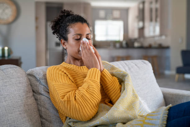 Cold sick african woman blowing nose Portrait of young black woman sneezing in to tissue at home. Sick african woman wrapped in blanket sitting on sofa blowing her nose at home. Ill girl sneezing with runny nose in winter. sinusitis photos stock pictures, royalty-free photos & images