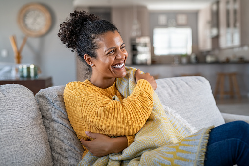 Beautiful black girl sitting on couch wrapped under blanket and laughing. Cheerful african american woman sensitive to the cold relaxing at home on sofa. Carefree and happy mid woman hugging self with warm blanket in winter.