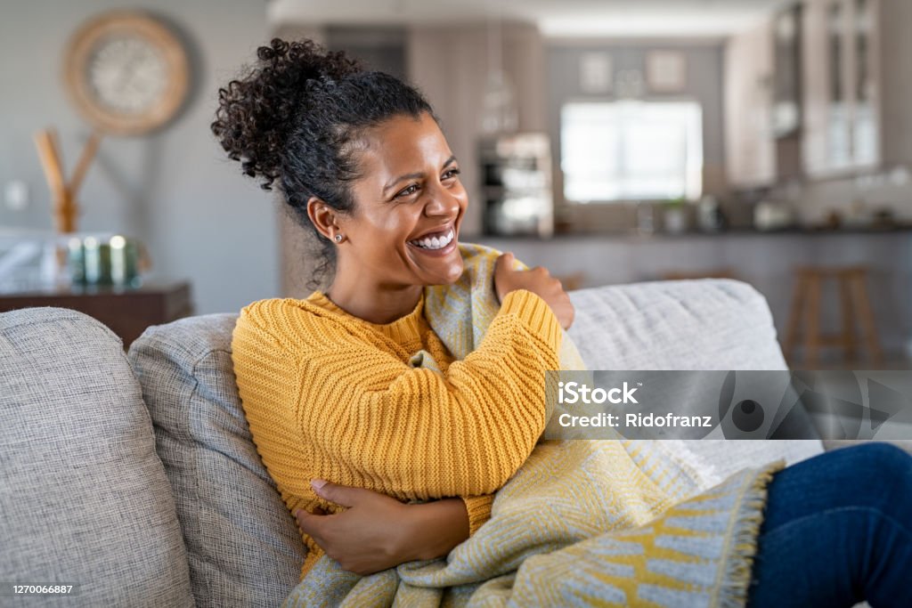 Joyful african woman with blanket on couch laughing Beautiful black girl sitting on couch wrapped under blanket and laughing. Cheerful african american woman sensitive to the cold relaxing at home on sofa. Carefree and happy mid woman hugging self with warm blanket in winter. Women Stock Photo