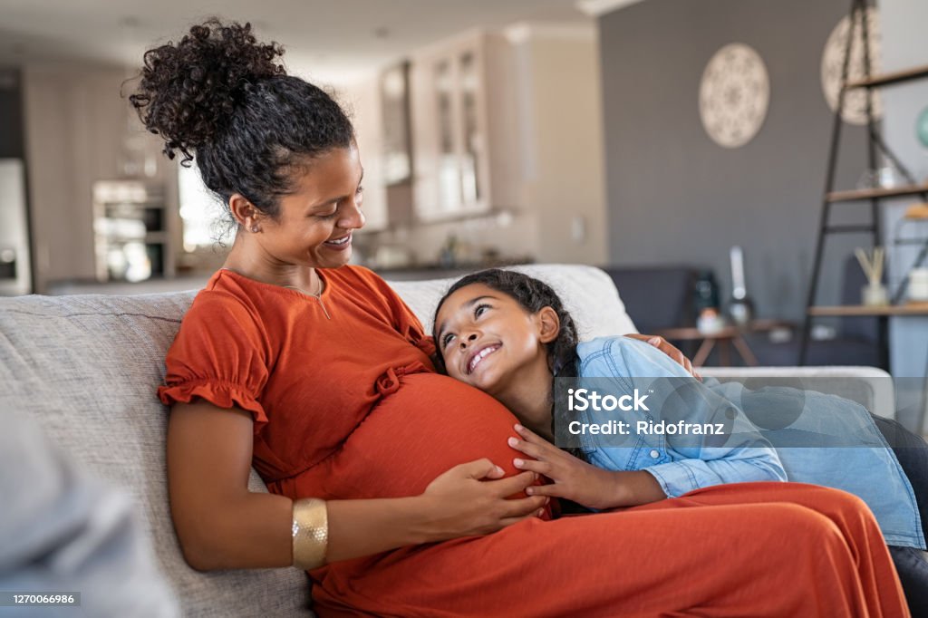 Happy lovely daughter hugging pregnant mother Happy mixed race daughter hugging belly of her expecting mother while relaxing on couch at home. African girl listening to baby movements while embracing pregnant woman. Pregnant black mom and future sister relaxing together on sofa at home. Pregnant Stock Photo