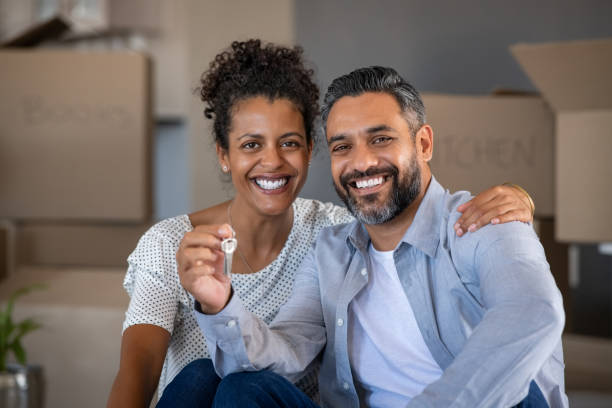 Mixed race couple holding new house keys Middle aged multiethnic couple embracing and holding house keys with carboard boxes behind them. Cheerful couple sitting on floor in new home. Portrait of indian man and african woman showing house keys while looking at camera after relocation. key package stock pictures, royalty-free photos & images