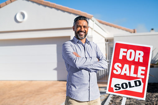 Portrait of confident indian man with crossed arms standing near sold signboard outside new home. Happy satisfied man excited to buy new home looking at camera. Successful mixed race real estate agent purchasing new home for investment purpose with copy space.