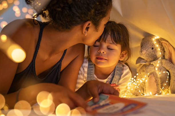 loving ethnic mother kissing daughter in cozy hut - child reading mother book imagens e fotografias de stock