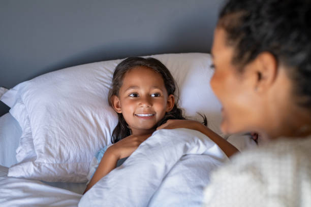 Little cute girl talking to mother in bed Cute smiling girl lying under blanket in bed wishing good night to mother. Happy lovely daughter talking with mom at bedtime. African mother putting indian child to sleep in comfortable bed. bedtime stock pictures, royalty-free photos & images