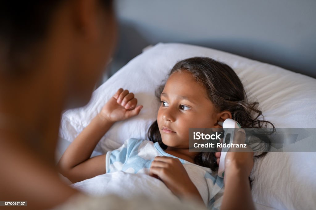 Mother checking daughter temperature with ear thermometer Close up of african mom using ear thermometer to check temperature of little sick girl lying on bed. Ill indian daughter resting in bed while woman checking fever. Portrait of girl with cold and flu symptoms while mother sitting next to hear. Child Stock Photo