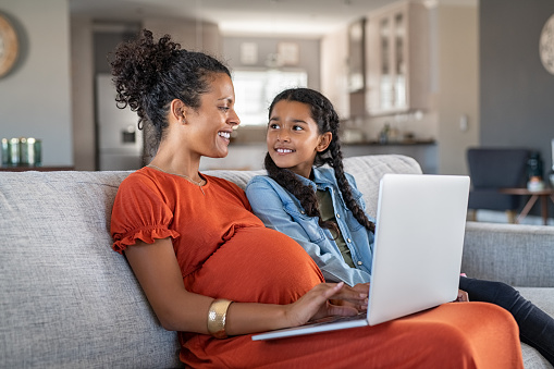 Pregnant black woman working on laptop while sitting on sofa and smiling to daughter. Cheerful expecting mother and girl relaxing on couch and using laptop to book vacation. Happy woman with baby bump and daughter working on computer from home during maternity leave.