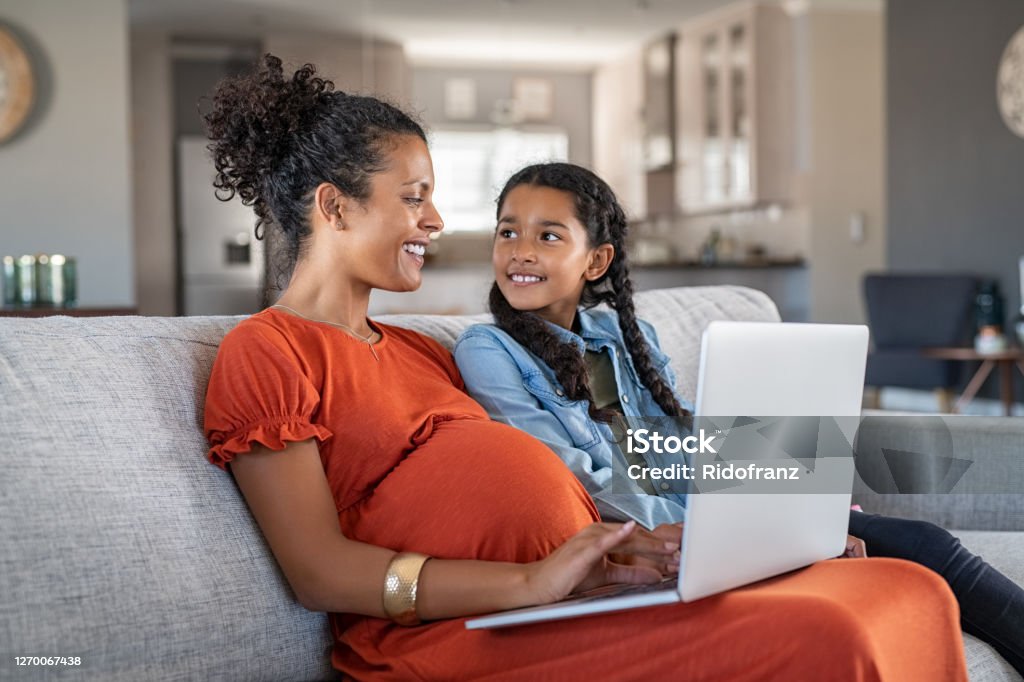 African pregnant woman working rom home in maternity leave Pregnant black woman working on laptop while sitting on sofa and smiling to daughter. Cheerful expecting mother and girl relaxing on couch and using laptop to book vacation. Happy woman with baby bump and daughter working on computer from home during maternity leave. Pregnant Stock Photo