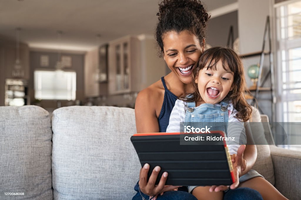 Ethnic mother and little girl having fun with digital tablet Young black mother and smiling daughter playing on digital tablet at home with copy space. Young african american woman with cheerful and excited little girl using digital tablet while relaxing on couch at home. Family Stock Photo