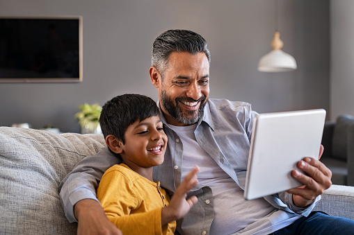 Indian father and smiling son sitting on couch using digital tablet at home. Man and boy using digital tablet for video calling at home while spending time together. Middle eastern dad with son doing videocall during quarantine.