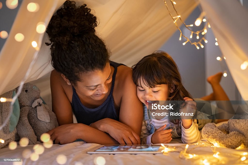 Mother and daughter using digital tablet inside illuminated cozy hut African mother and cute smiling girl using digital tablet while lying in illuminted tent in kid bedroom. Cheerful ethnic woman and lovely daughter on video call under a cozy hut. Lovely little girl with mom watching cartoon on digital tablet in bedroom. Family Stock Photo
