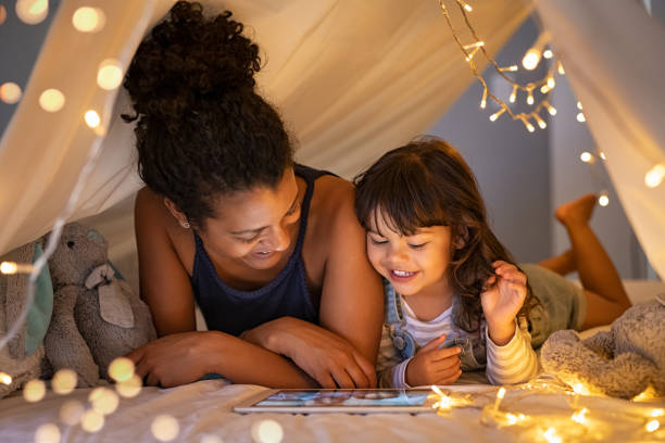 Mother and daughter using digital tablet inside illuminated cozy hut African mother and cute smiling girl using digital tablet while lying in illuminted tent in kid bedroom. Cheerful ethnic woman and lovely daughter on video call under a cozy hut. Lovely little girl with mom watching cartoon on digital tablet in bedroom. hut stock pictures, royalty-free photos & images