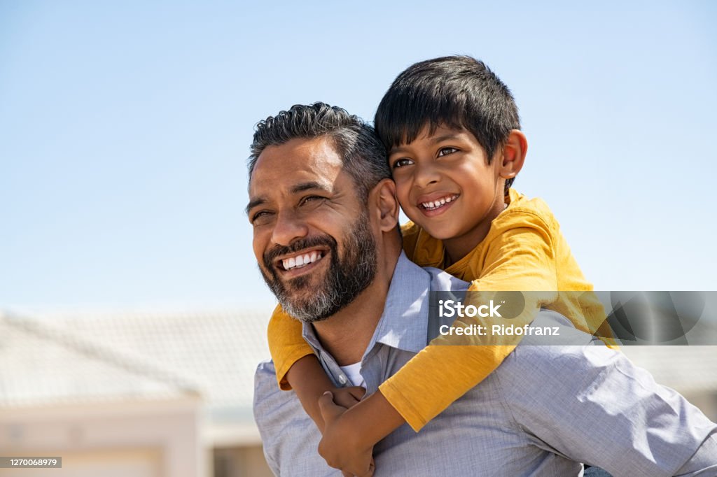 Happy smiling indian father giving son ride on back Happy middle eastern child enjoying ride on father back outdoor. Smiling young dad giving piggyback ride to son on street while looking away with copy space. Latin cheerful man carrying on shoulder indian kid: future and vision concept . Father Stock Photo
