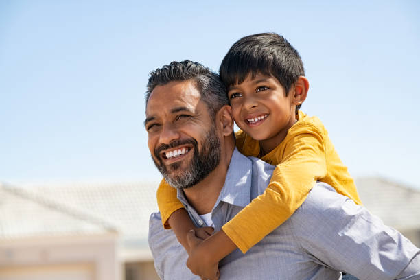 Happy smiling indian father giving son ride on back Happy middle eastern child enjoying ride on father back outdoor. Smiling young dad giving piggyback ride to son on street while looking away with copy space. Latin cheerful man carrying on shoulder indian kid: future and vision concept . hispanic stock pictures, royalty-free photos & images