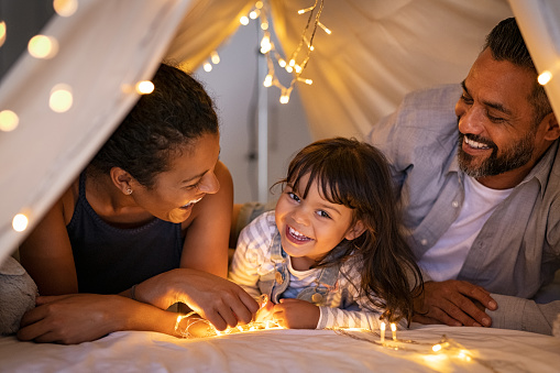 Happy latin daughter with african mom and indian dad playing in kid tent at home. Little girl talking to mother and father lying on bed in illuminated hut. Funny cute girl playing with multiethnic parent in beautiful cozy shelter at home.