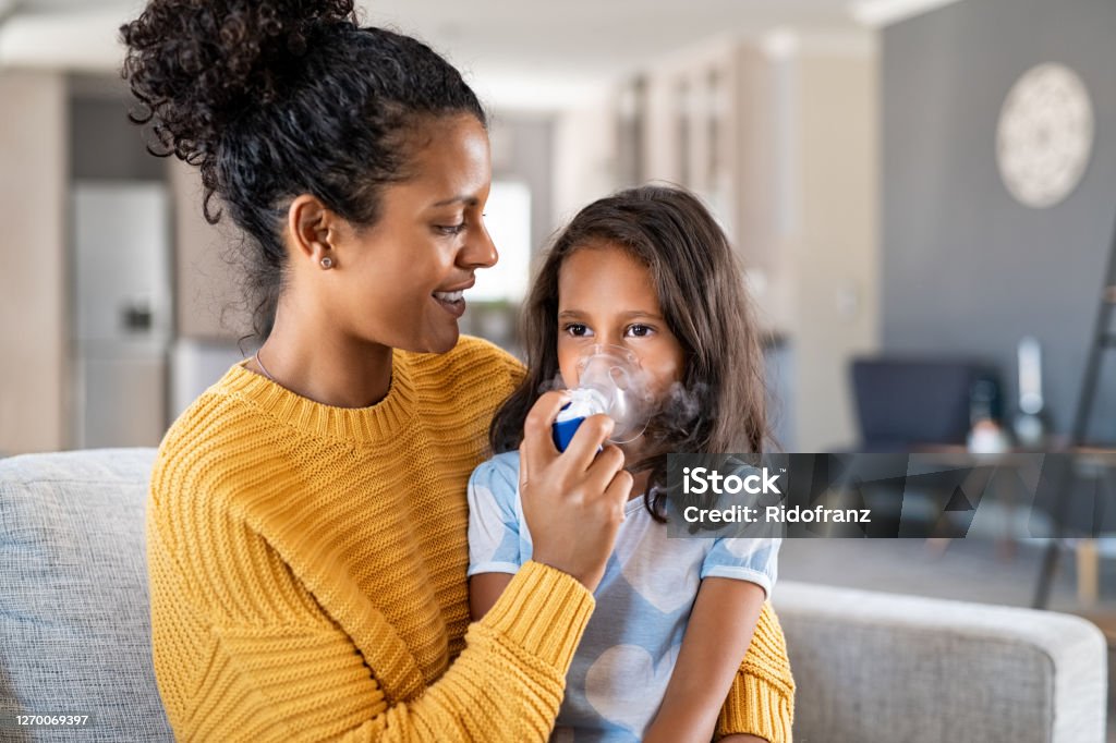 African mother helping child use nebulizer aerosol Little indian girl making inhalation with nebulizer at home with lovely mother. Woman makes inhalation to a sick child while embracing her. Mom helping daughter with cold and flu to inahale nebuliser aerosol sitting on couch. Asthmatic Stock Photo