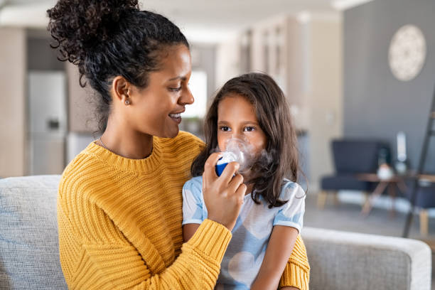 African mother helping child use nebulizer aerosol Little indian girl making inhalation with nebulizer at home with lovely mother. Woman makes inhalation to a sick child while embracing her. Mom helping daughter with cold and flu to inahale nebuliser aerosol sitting on couch. asthmatic stock pictures, royalty-free photos & images