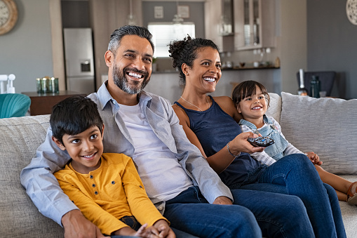 Happy indian man and african woman relaxing with daughter and son watching television at home together. Cheerful ethnic family relaxing on sofa at home watching movie with children. Woman controlling remote while looking at cartoons with middle eastern husband and kids.