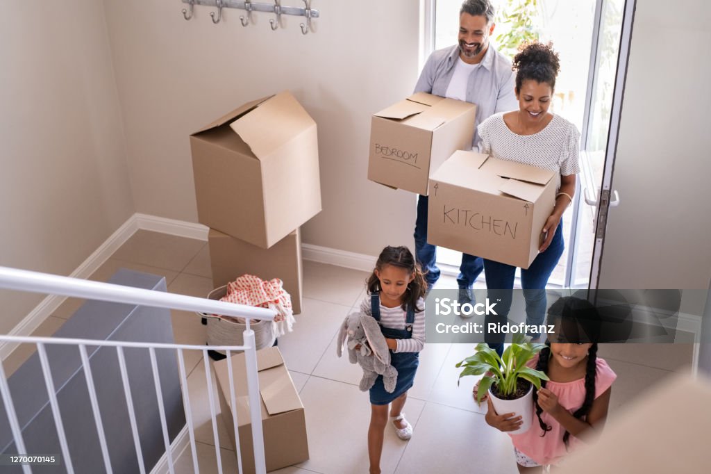 Multiethnic family moving in new home Ethnic family with two children carrying boxes and plant in new home on moving day. High angle view of happy smiling daughters helping mother and father with cardboard boxes in new house. Top view of excited kids having fun walking up stairs running to their rooms while parents holding boxes. Moving House Stock Photo