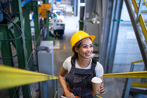Female engineer in uniform and hardhat climbing metal industrial steps of factory building. Holding cup of coffee and smiling.