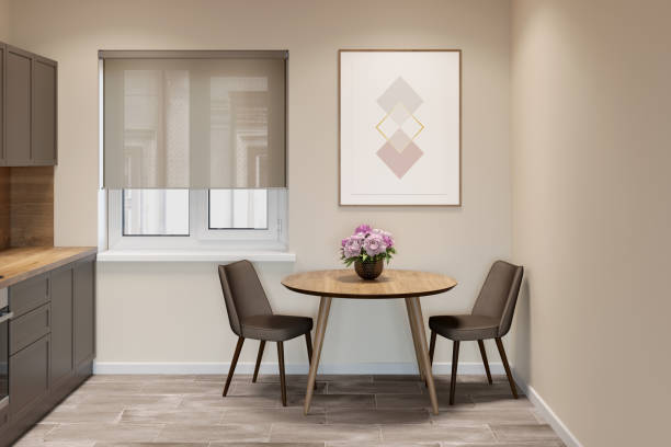 Beige kitchen interior with a window with blinds between a round table with two chairs and kitchen cabinets. There is a vertical poster on the wall above the table. Front view.