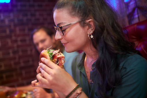 Close-up of hispanic woman holding delicious burger and smelling it in retro restaurant