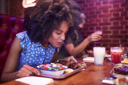 Close-up of young african-american woman with afro hair enjoying and smelling bbq ribs in retro restaurant with friends
