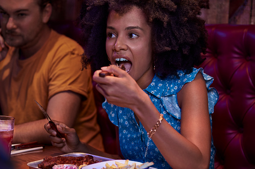 African-american woman with afro eating ribs with fork and knife in rustic restaurant with friends