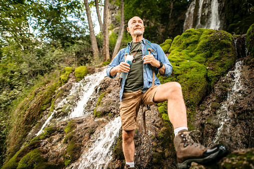 Senior Backpacker Hiker Standing in a front of a Mountain Waterfall, holding Water Bottle, Enjoying the View