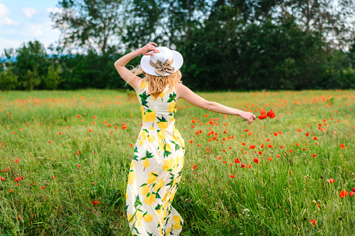 Blonde in a dress and hat runs with joy across the poppy field