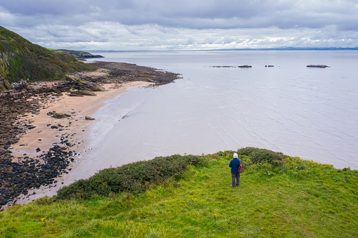 One senior man standing on the edge of a cliff next to the coastline of Dumfries and Galloway, south west Scotland. Although it is a summer morning the sky is overcast with grey clouds.