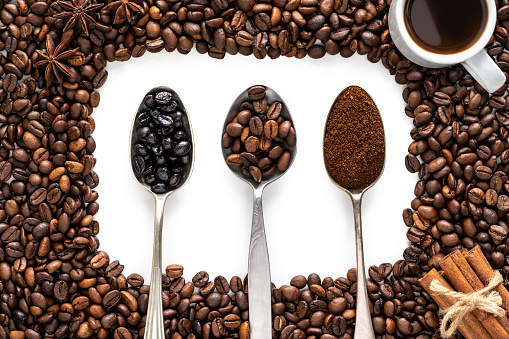 Coffe beans with two toast levels and ground coffee on three spoons and coffee cup in a border of coffee beans over white