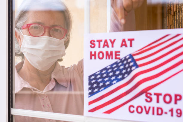 una donna anziana dietro la finestra indica il messaggio di rimanere a casa indossando una maschera chirurgica per evitare il contagio del coronavirus covid-19 - concetto di speranza per il futuro - isolated despair hope assistance foto e immagini stock