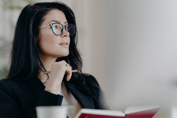 istock Thoughtful European woman makes notes in notepad, holds pen and writes down some ideas, makes list of goals, plans day in personal organizer, wears transparent glasses, thinks about something 1270100192