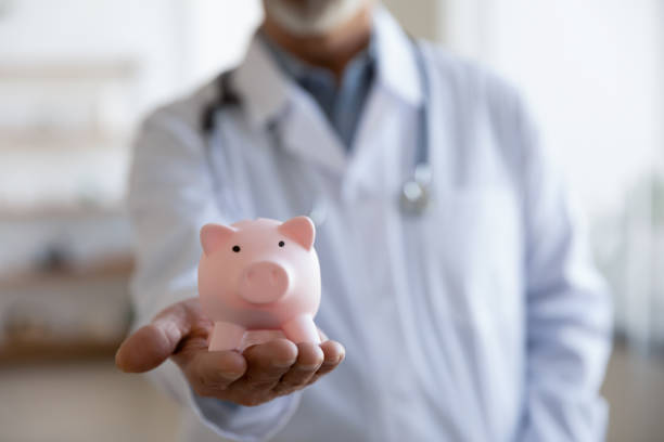close up mature doctor wearing uniform holding pink piggy bank - currency stethoscope medicare usa imagens e fotografias de stock
