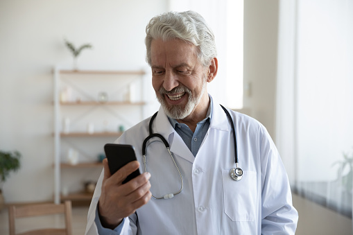 Smiling mature doctor wearing white uniform coat with stethoscope looking at smartphone screen, making video call, friendly senior therapist gp consulting patient online, telemedicine concept
