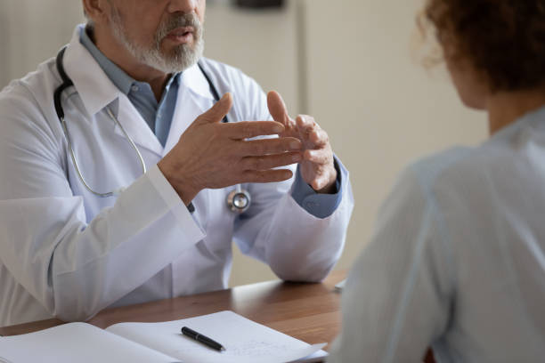 Close up serious senior doctor talking to female patient Close up serious senior doctor talking to female patient at meeting in hospital, mature physician gp consulting woman about treatment, explaining, discussing medical checkup results, sitting at desk general view stock pictures, royalty-free photos & images