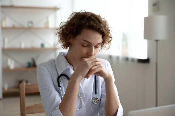 Close up thoughtful woman doctor sitting at work desk Close up thoughtful woman doctor with closed eyes sitting at work desk in hospital, meditating at workplace, female therapist physician gp wearing white coat uniform praying, asking help melancholy computer support specialist stock pictures, royalty-free photos & images
