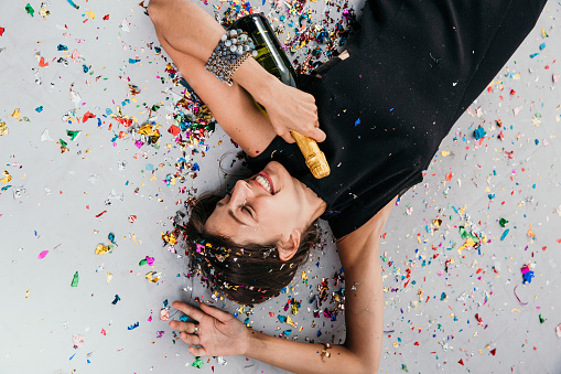 Satisfied happy rich lady lying on the floor full of confetti, holding a Champagne bottle, and celebrating new moments. A productive and successful concept