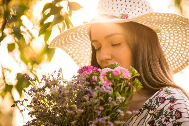 Close up of young woman's face smelling a bunch of flowers recently cut during springtime. She wears a hat.