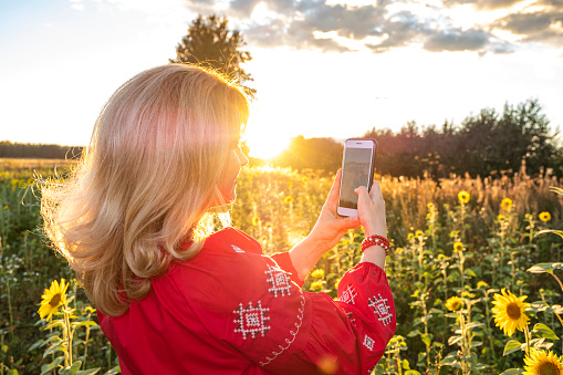 Woman 40 years old, Caucasian, blonde. Posing and taking a selfie on a smartphone. Woman in a red dress. Against the background of Sunset and sunflowers floating. Back view.