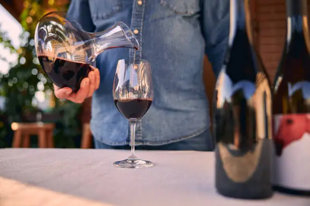 Cropped photo of a glass on the table and a man in denim shirt pouring red wine into it from the decanter