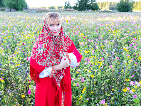 Young Caucasian woman, traditional national clothes of Russia and Ukraine, red sundress and shawl on her head. Against the background of meadows with wildflowers. Portrait.