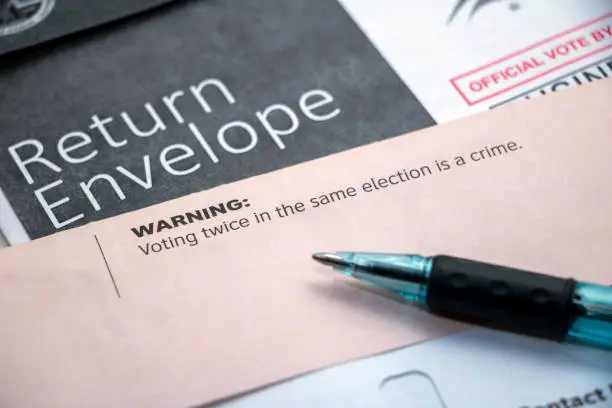 Closeup on pile of voter mail with a pen laying on top, focused on the top of the envelope with text, 'Warning: Voting twice in the same election is a crime."